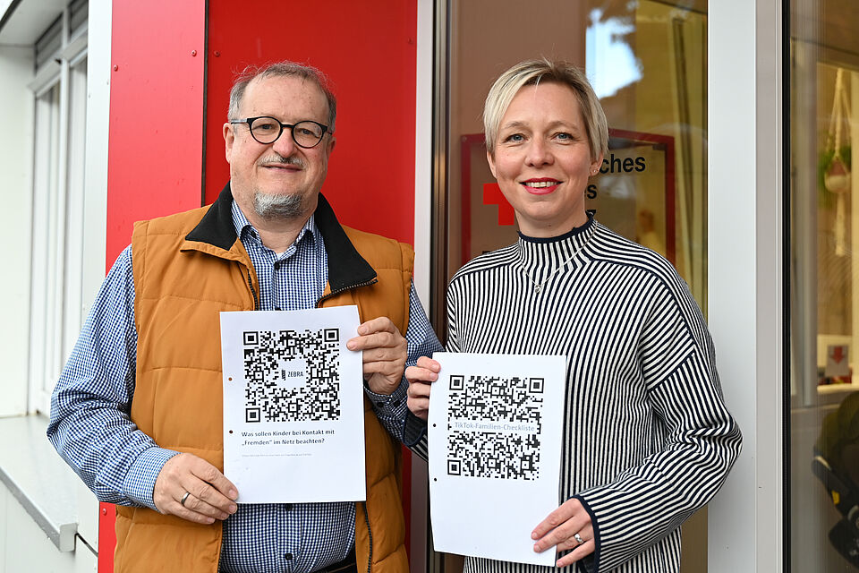 Zwei Personen stehen vor einem Gebäude mit rotem Element und einem DRK-Logo im Hintergrund. Sie halten jeweils ein Blatt Papier mit großen QR-Codes in die Kamera. Die linke Person trägt eine Brille, ein kariertes Hemd und eine braune Weste, die rechte Person trägt einen weiß-dunkelblau gestreiften Pullover. Beide lächeln und präsentieren die Ausdrucke frontal.