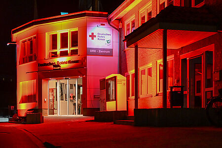 Nachts rot angestrahltes DRK-Gebäude mit erleuchtetem Schild „Deutsches Rotes Kreuz – DRK-Zentrum“