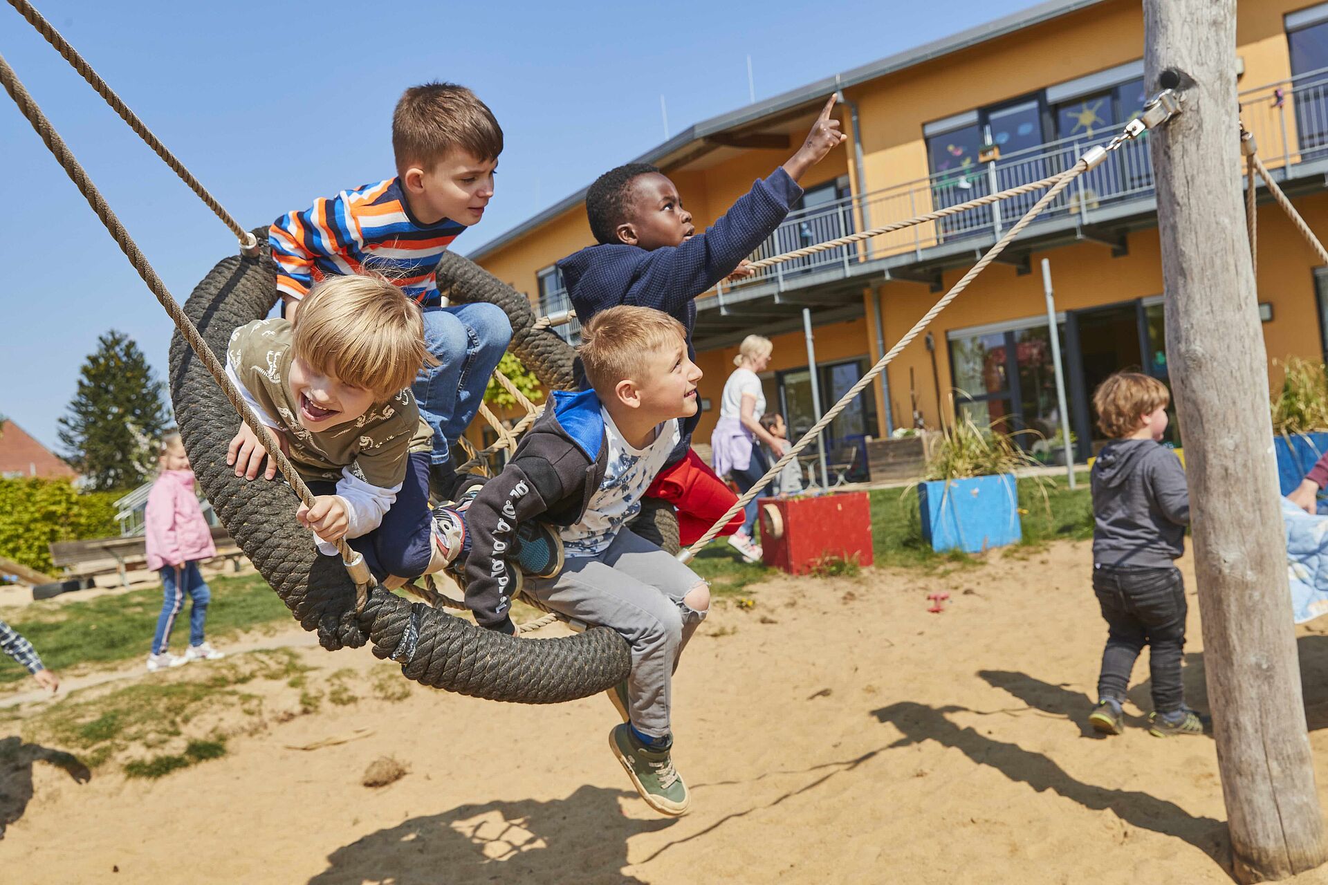 Mehrere Kinder spielen auf einem Reifen-Schaukel auf einem Sandspielplatz vor einem Gebäude.