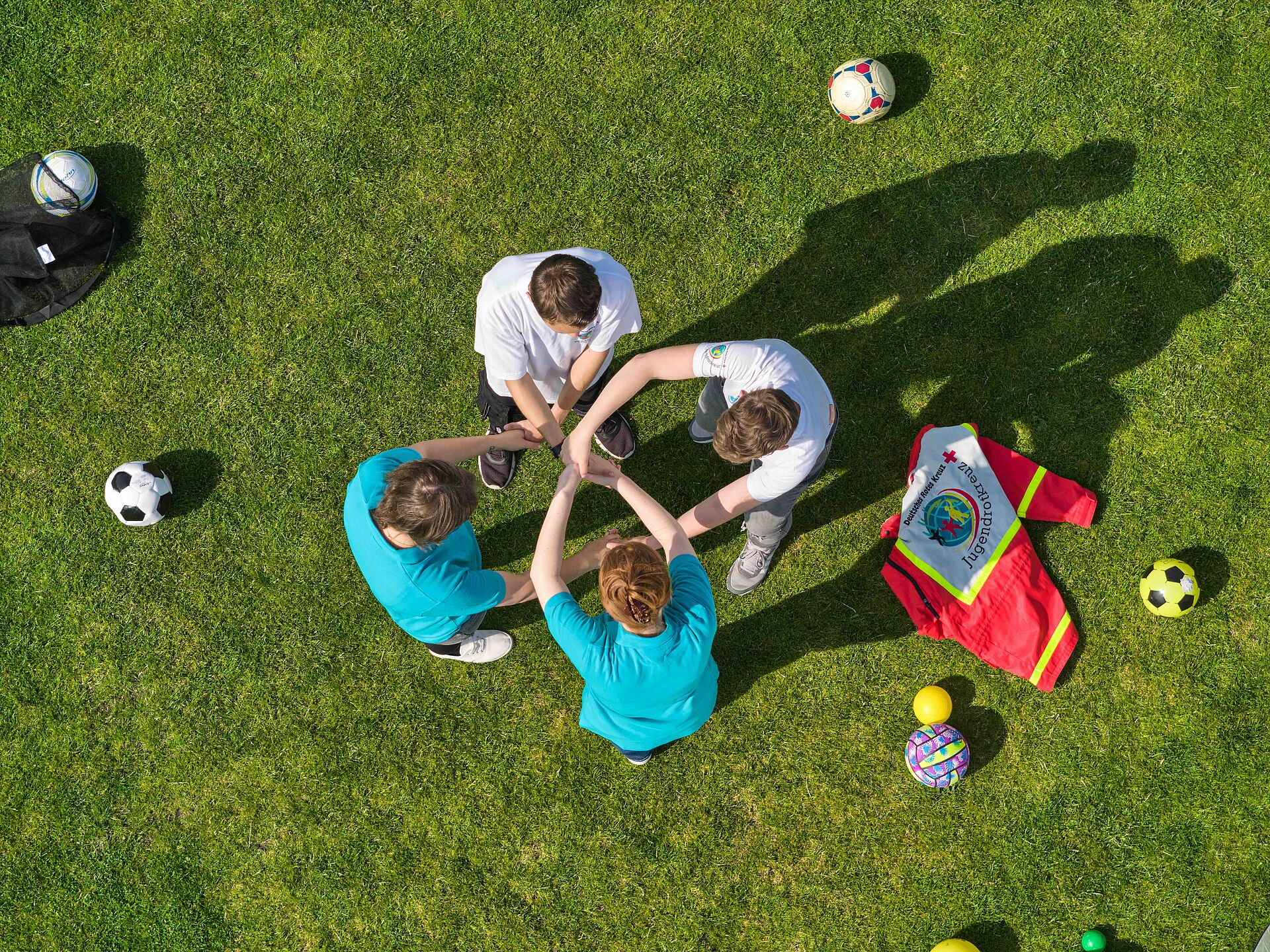Vier Personen stehen im Kreis auf einem grünen Rasenplatz, legen ihre Hände übereinander in die Mitte und zeigen so Teamgeist. Um sie herum liegen verschiedene Sportbälle und eine rote Tasche des Jugendrotkreuzes.