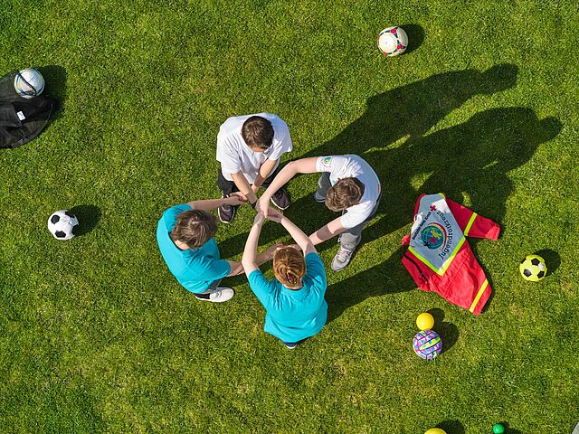 Vier Personen stehen im Kreis auf einem grünen Rasenplatz, legen ihre Hände übereinander in die Mitte und zeigen so Teamgeist. Um sie herum liegen verschiedene Sportbälle und eine rote Tasche des Jugendrotkreuzes.