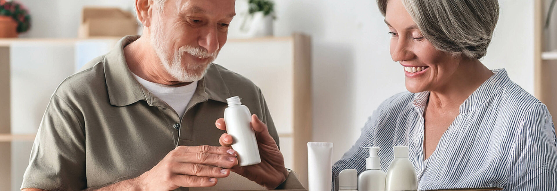 Ein lächelndes älteres Paar begutachtet gemeinsam Produkte aus einer geöffneten Box, der Mann hält eine weiße Flasche in der Hand.
