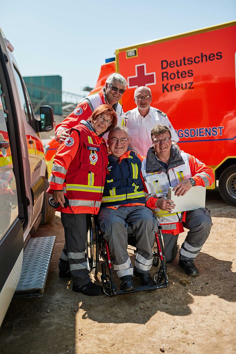 Lageplan Fünf lächelnde DRK-Helfer in Uniformen, einer davon im Rollstuhl, posieren vor einem roten Rettungswagen mit dem Schriftzug "Deutsches Rotes Kreuz".