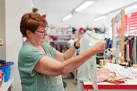 Kleidershop Eine Mitarbeiterin des Kleidershops hält ein weißes Babybody mit blauem Aufdruck in den Händen und begutachtet es aufmerksam. Im Hintergrund sind Regale mit verschiedenen Waren und Kleidungsstücken zu sehen.
