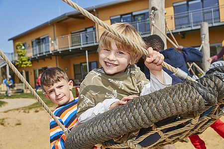 Zwei Kinder spielen fröhlich auf einem Seilnetz-Klettergerüst im Außenbereich einer Kita. Im Hintergrund ist das moderne Gebäude der Einrichtung mit Balkonen und großen Fenstern zu sehen. Die Kinder wirken konzentriert und glücklich, während sie das Klettergerüst erkunden. Die Szene vermittelt eine lebendige und kindgerechte Atmosphäre.