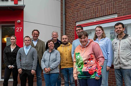 Mitglieder des Klientenbeirats posieren gemeinsam vor einem Backsteingebäude mit der Hausnummer 27. Die Gruppe ist in zwei Reihen aufgestellt und lächelt in die Kamera; die Stimmung ist freundlich und locker.