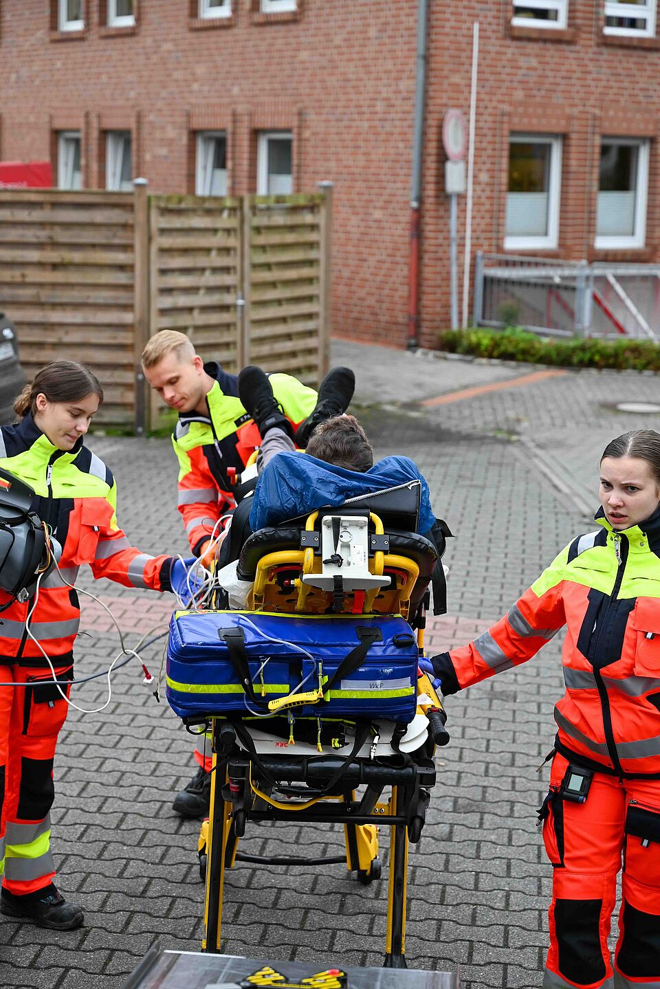 Mehrere Rettungskräfte in orange-gelber Einsatzkleidung transportieren eine Person auf einer Trage. Die Trage ist mit medizinischem Equipment ausgestattet, im Hintergrund stehen Backsteingebäude.