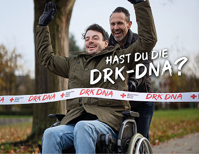 Ein lachender Mann im Rollstuhl mit erhobenen Armen fährt durch ein Zielband mit der Aufschrift "DRK DNA", geschoben von einem anderen lachenden Mann.