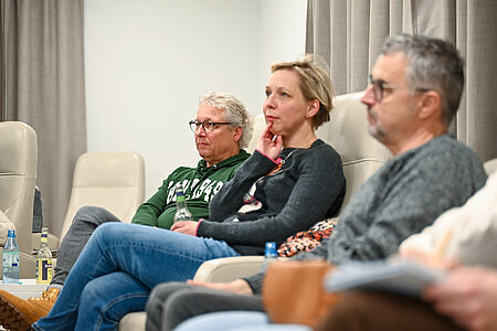 Mehrere Erwachsene sitzen in einem hellen Raum auf gepolsterten Stühlen und hören aufmerksam zu. Sie blicken nach vorn, wirken konzentriert und nachdenklich. Vor ihnen stehen Getränke auf dem Boden und Tischen. Die Szene erinnert an eine Gesprächsrunde oder ein Informations­treffen.
