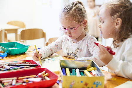 DRK-Kita "Abenteuerland" Zwei Kinder sitzen an einem Tisch und malen gemeinsam. Das Kind im Vordergrund trägt eine Brille und ein weißes T-Shirt mit einem Motiv. Auf dem Tisch liegen bunte Stifte in einer Schachtel sowie ein grünes Gefäß. Im Hintergrund sind weitere Kinder und Stühle zu erkennen.
