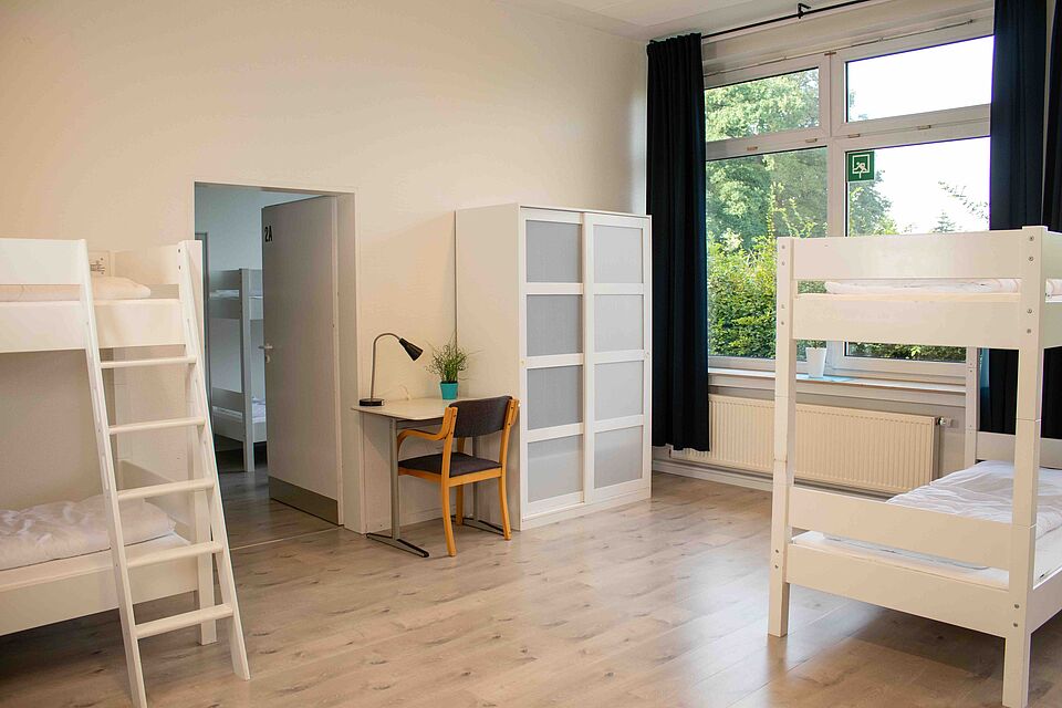 Innenraum mit hellen Wänden und Boden. Es sind weiße Etagenbetten, ein Schreibtisch mit Lampe und Stuhl sowie ein Schrank zu sehen.