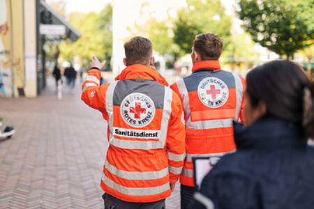 Zwei Personen in orangefarbenen Warnwesten mit der Aufschrift 'Sanitätsdienst' und dem Logo des Deutschen Roten Kreuzes (DRK) von hinten fotografiert. Sie tragen Rucksäcke und gehen auf einem Gehweg in einer städtischen Umgebung.