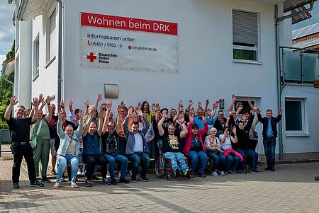 Vor einem weißen Gebäude mit dem Schild „Wohnen beim DRK“ sitzt und steht eine große Gruppe von rund 35 Menschen. Sie sind gemischt aus Männern und Frauen unterschiedlichen Alters. Einige sitzen auf Bänken, eine Person im Rollstuhl ist ebenfalls sichtbar. Alle heben beide Arme fröhlich in die Luft und lächeln in die Kamera. Am Gebäude hängt ein großes rotes Schild mit der Aufschrift „Wohnen beim DRK“ sowie Kontaktinformationen des Deutschen Roten Kreuzes.