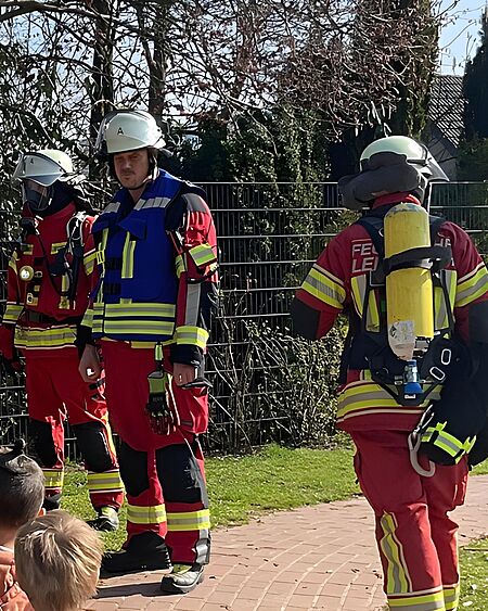 Drei Feuerwehrleute in voller Schutzausrüstung mit Helmen und reflektierenden Streifen stehen im Freien. Ein Feuerwehrmann trägt einen Pressluftatmer auf dem Rücken, während im Hintergrund Kinder zu sehen sind.