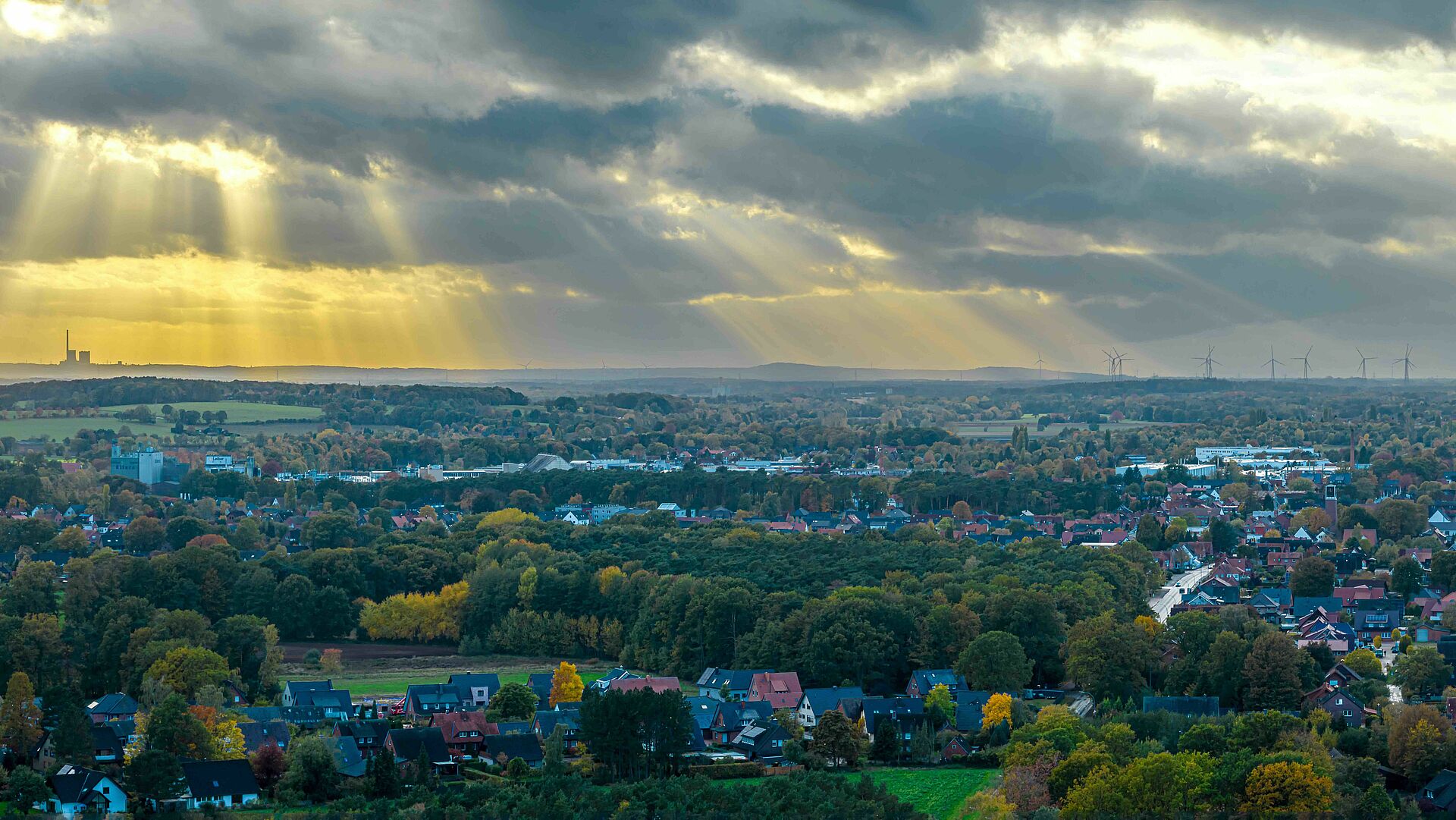 Ein Luftbild zeigt eine weite Landschaft des Tecklenburger Landes mit Häusern, Bäumen und Feldern, über denen Sonnenstrahlen durch die Wolken brechen. Am Horizont sind Industriebauten und Windräder zu erkennen.