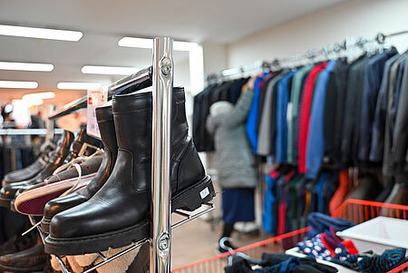 In einem Secondhand- oder Sozialkaufhaus stehen vorne schwarze Lederstiefel und weitere Schuhe ordentlich auf einem Metallregal. Im Hintergrund hängt eine Vielzahl von Jacken und Kleidung auf einem Kleiderständer, während eine Person in heller Jacke danach greift. Die Szene wirkt wie ein sortierter Verkaufs- oder Ausgabebereich.