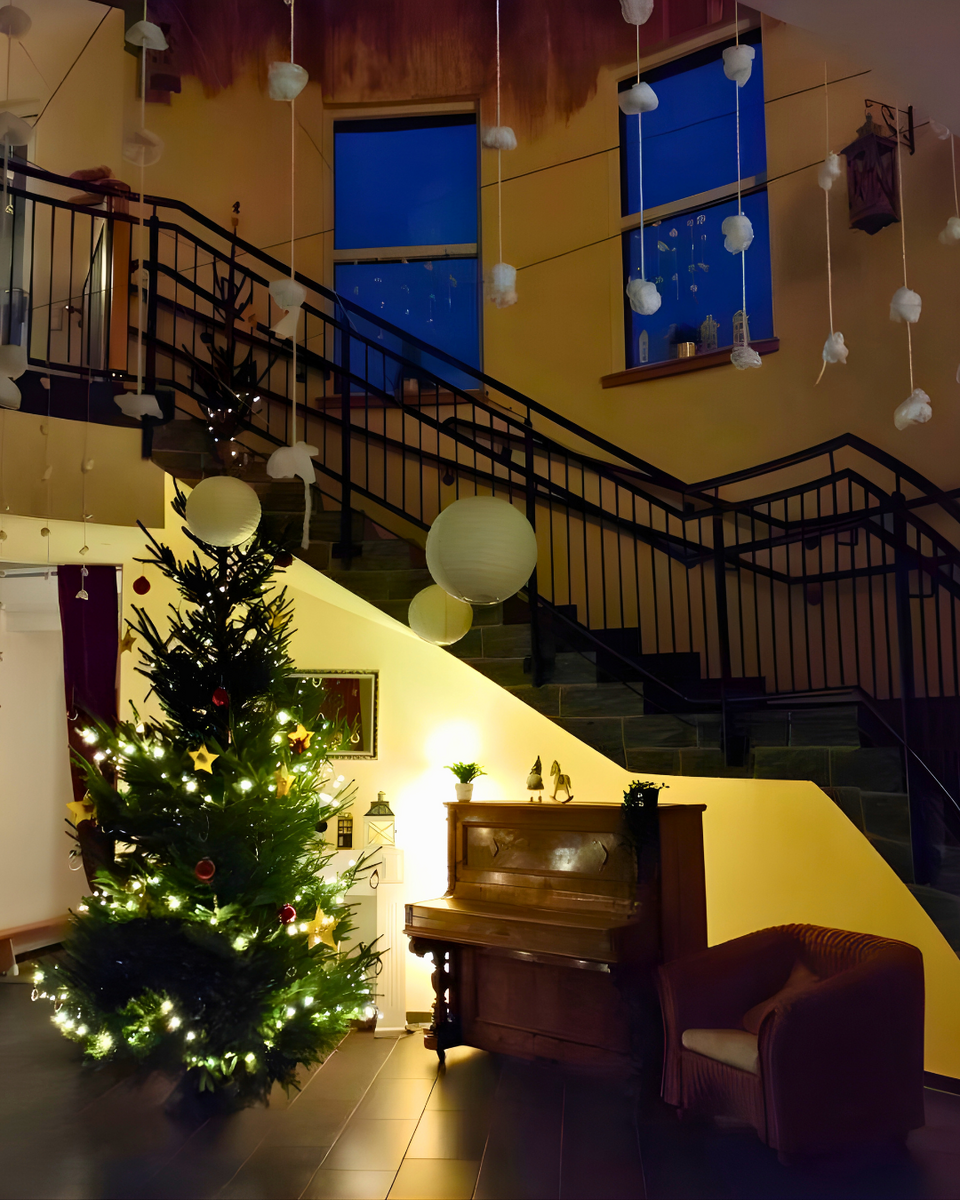 Ein festlich geschmückter Eingangsbereich mit einem beleuchteten Weihnachtsbaum links, daneben ein altes Klavier und ein Sessel; darüber führen Treppen nach oben, von der Decke hängen weiße Deko-Elemente und Papierlaternen, und durch die Fenster ist der dunkelblaue Abendhimmel zu sehen.