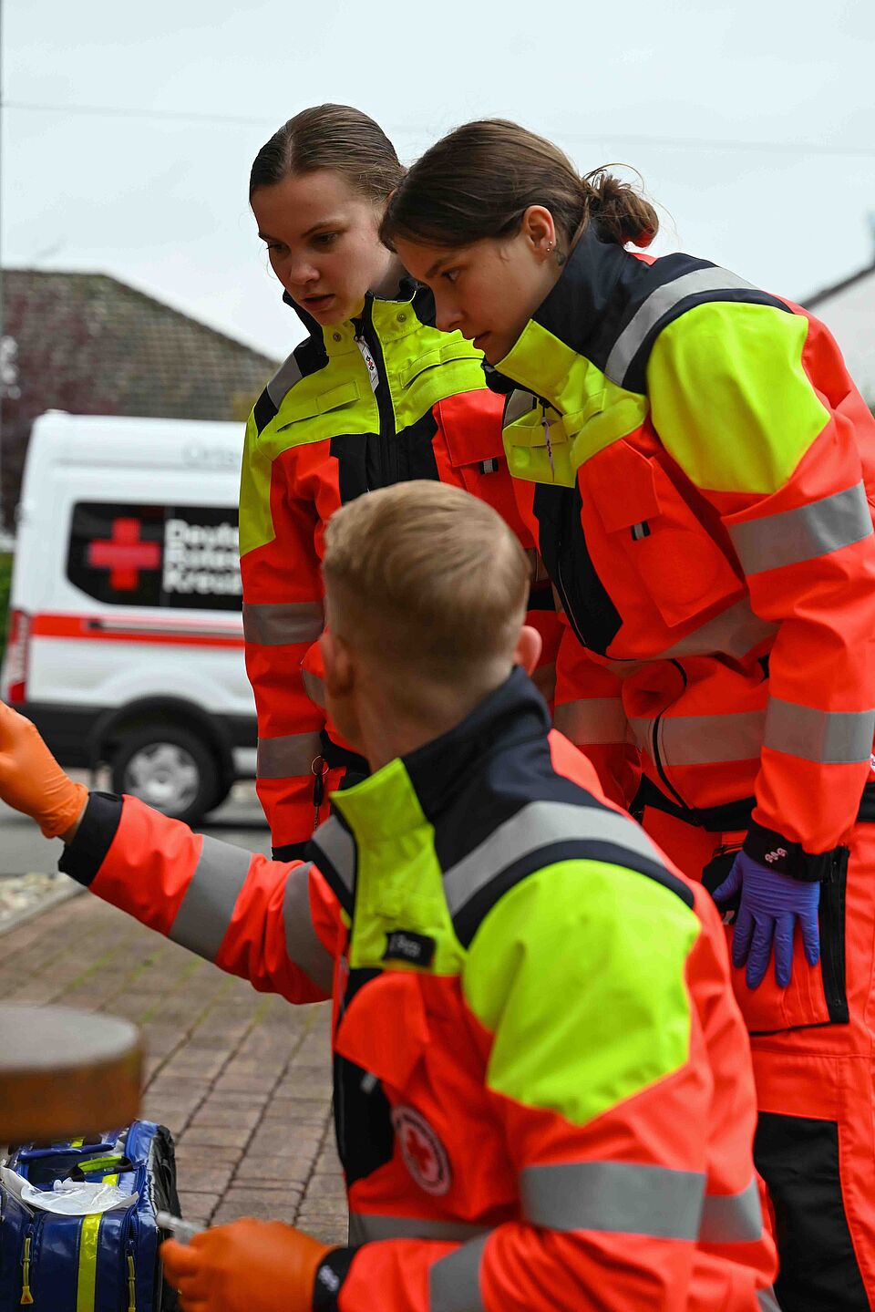 Zwei Rettungskräfte in orange-gelber Einsatzkleidung beugen sich zu einem Kollegen, der kniet und mit Handschuhen arbeitet. Im Hintergrund ist ein Einsatzfahrzeug des DRK zu sehen.