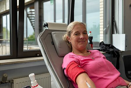Eine lächelnde blonde Frau liegt in einem Stuhl und spendet Blut, ihr Arm ist mit einem roten Verband fixiert.