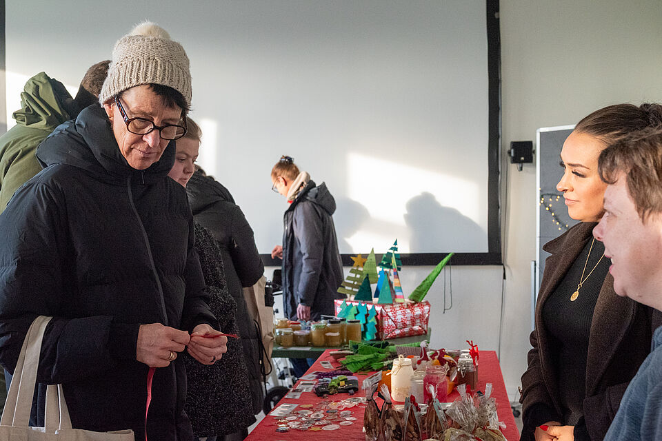 Mehrere Menschen stehen an einem Tisch mit handgemachten Weihnachtsartikeln. Im Vordergrund betrachtet eine ältere Person mit Mütze und Brille kleine Bastelarbeiten, während zwei weitere Personen hinter dem Tisch stehen und ins Gespräch vertieft sind. Auf dem Tisch liegen dekorative Gläser, Kerzen, Karten und kleine Weihnachtsbäume. Die Szene spielt in einem hellen Innenraum und vermittelt eine ruhige, vorweihnachtliche Marktatmosphäre.