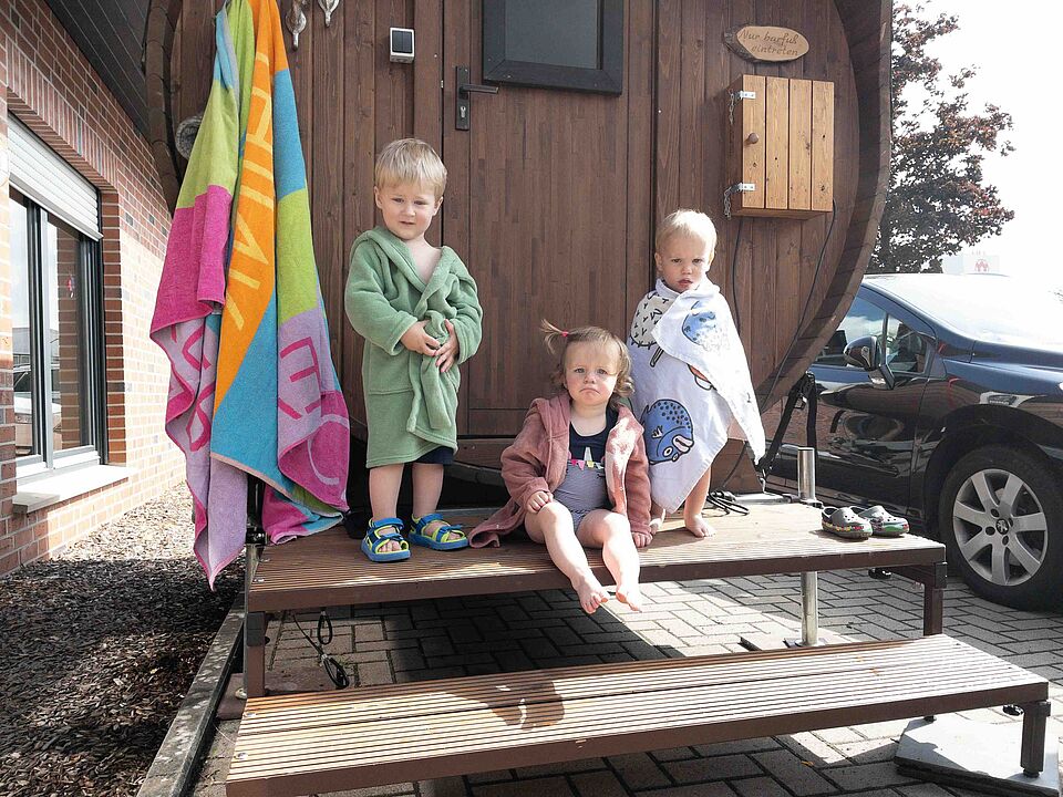 Drei kleine Kinder sitzen vor der braunen Fasssauna. 