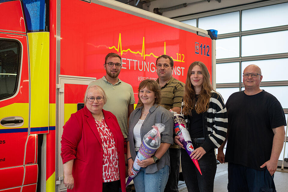 Sechs Personen stehen vor einem Rettungswagen mit roter und gelber Lackierung und der Aufschrift „Rettungsdienst 112, Kreis Steinfurt“. Zwei Frauen in der Mitte halten bunte Schultüten in den Händen, eine davon mit einem Plüsch-Pferd. Alle lächeln in die Kamera