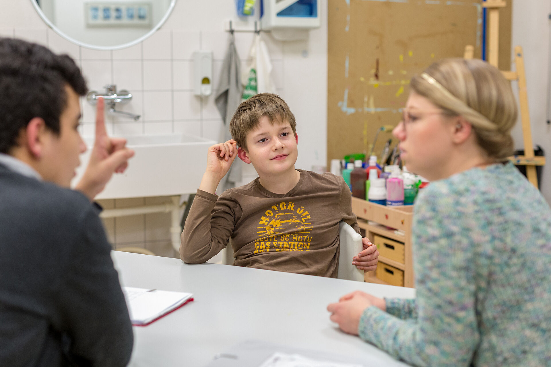 Ein lächelnder Junge sitzt an einem Tisch und schaut zu einer Frau mit Brille und blondem Haarband, während ein weiterer Mann mit dunklen Haaren und dunklem Hemd im Vordergrund mit erhobenem Zeigefinger zu sehen ist.