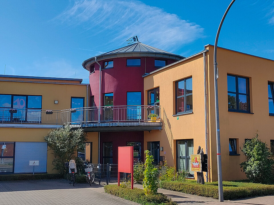 Modernes, zweigeschossiges Gebäude mit gelber und roter Fassade, großen Fenstern und einem runden Turm mit silbernem Dach. Vor dem Eingang stehen Fahrräder, Pflanzen und ein rotes Schild. Rechts am Gebäude ist ein buntes Logo mit einer Figur und dem Schriftzug „Kita Biberburg“ zu sehen. Der Himmel ist blau mit wenigen Wolken.