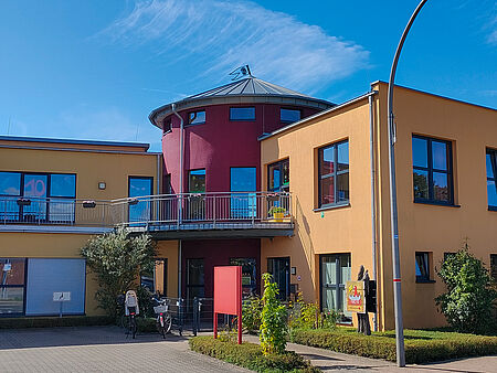 Modernes, zweigeschossiges Gebäude mit gelber und roter Fassade, großen Fenstern und einem runden Turm mit silbernem Dach. Vor dem Eingang stehen Fahrräder, Pflanzen und ein rotes Schild. Rechts am Gebäude ist ein buntes Logo mit einer Figur und dem Schriftzug „Kita Biberburg“ zu sehen. Der Himmel ist blau mit wenigen Wolken.