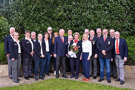 Gruppe von fünfzehn Personen in Uniformen des Deutschen Roten Kreuzes steht lächelnd in zwei Reihen vor einem grünen Buschhintergrund. Die Personen tragen dunkle Jacken, weiße Hemden und teilweise rote Krawatten oder Schals. Eine Person in der Mitte hält einen Blumenstrauß mit roten und rosafarbenen Rosen.