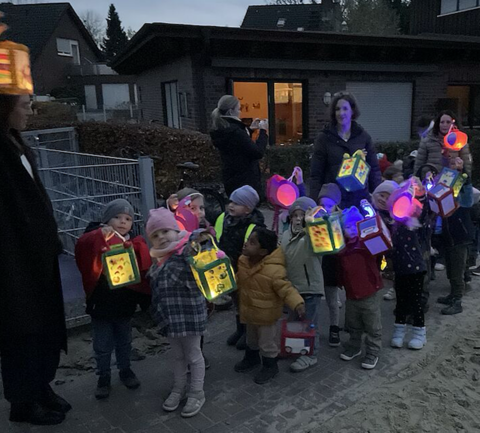 Eine Gruppe von Kita-Kindern steht in der Dämmerung auf dem Außengelände. Sie halten bunte, leuchtende Laternen in den Händen. Mehrere erwachsene Begleitpersonen stehen im Hintergrund. Die Kinder tragen warme Herbstkleidung, und die Laternen erhellen ihre Umgebung. Hinter ihnen sind Gebäude und ein Spielplatzbereich zu sehen.