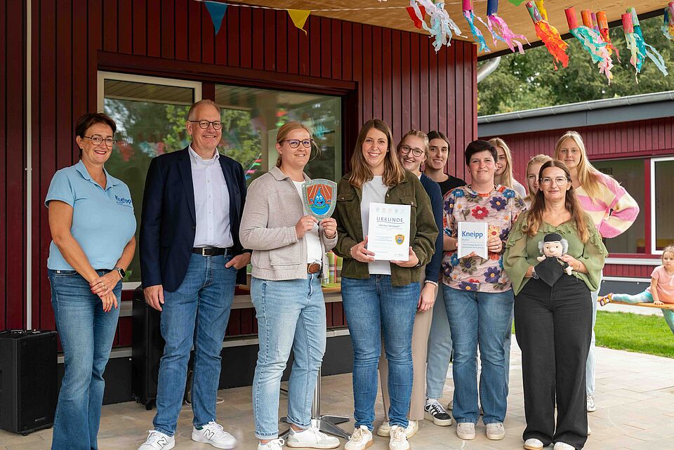 Eine Gruppe von neun Personen steht vor einem roten Holzgebäude, das mit bunten Girlanden und Papptellern geschmückt ist. Mehrere Personen halten Urkunden oder Bücher, darunter eines mit der Aufschrift „Kneipp Bund“. Eine Person hält eine Puppe im Arm. Die Gruppe wirkt fröhlich und feierlich, im Hintergrund sind weitere Gebäude und ein Spielplatz zu erkennen.