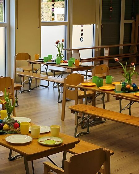 Gruppenraum mit mehreren Holztischen, die für eine Frühlingstafel oder Osterfeier gedeckt sind. Die Tische sind mit bunten Tellern, Bechern in Gelb und Grün sowie kleinen Blumenarrangements und bemalten Ostereiern dekoriert. Im Hintergrund sind große Fenster zu sehen, die viel Tageslicht hereinlassen.