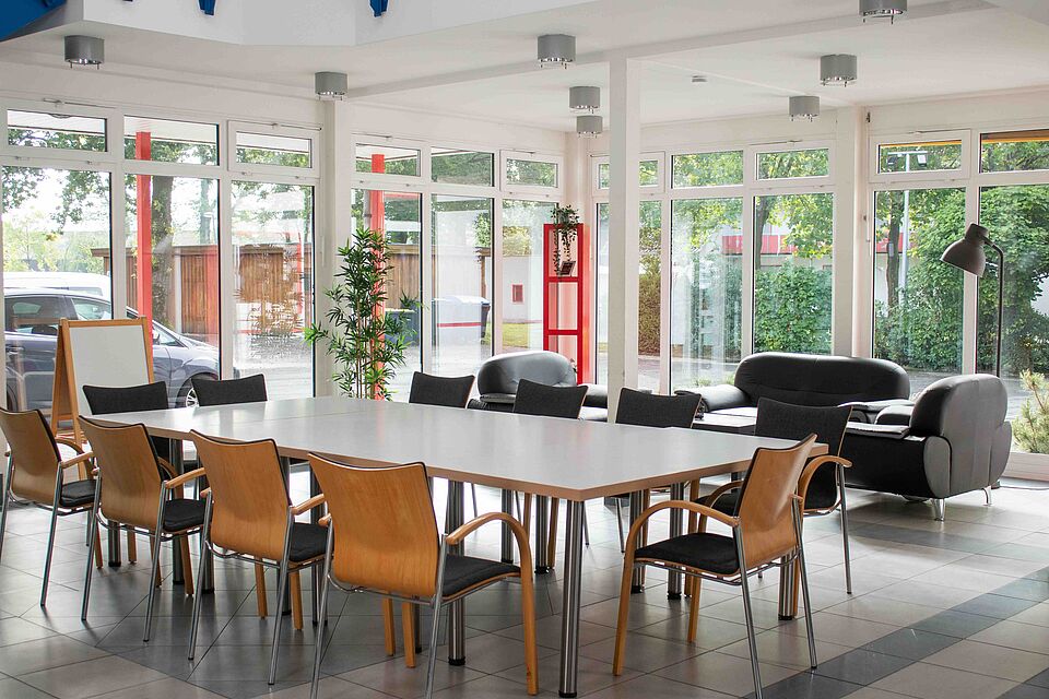 Ein heller Aufenthaltsraum mit großen Fenstern, Tischen und Stühlen. Im Hintergrund stehen Sofas und Pflanzen.