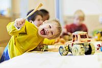 Ein lachendes Kind in einem gelben Malkittel hält einen Pinsel hoch, während es am Tisch mit Spielzeugtraktoren malt.