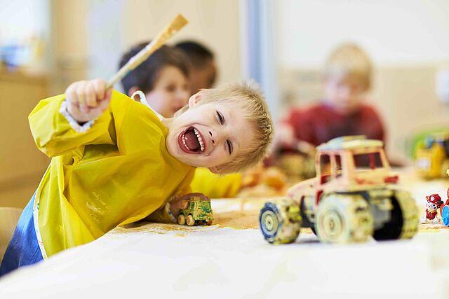 Ein lachendes Kind in einem gelben Malkittel hält einen Pinsel hoch, während es am Tisch mit Spielzeugtraktoren malt.