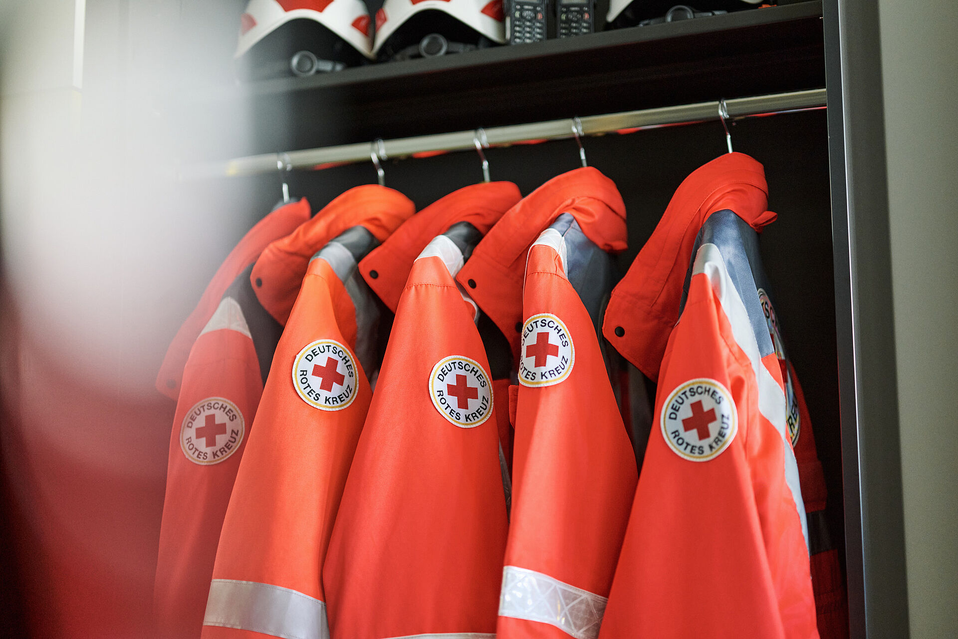 Mehrere orangefarbene Einsatzjacken des Deutschen Roten Kreuzes mit dem Logo und der Aufschrift hängen nebeneinander an Kleiderbügeln in einem Schrank, darüber liegen weiße Schutzhelme auf einem Regalbrett.