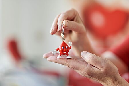 Eine Hand hält einen Schlüsselanhänger in Form eines lächelnden, roten Bluttropfens mit Armen und Beinen, der als Symbol für Blutspende und Engagement dient.