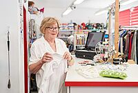 Eine Frau mit Brille hält ein weißes Kleidungsstück in einem Kleiderladen vor einem Verkaufstresen.
