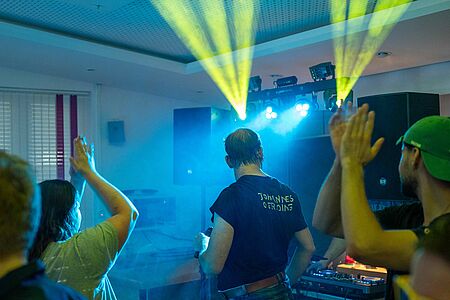Das Bild zeigt eine Diskoveranstaltung mit bunten Lichteffekten, bei der ein DJ Musik auflegt und Feiernde tanzen.