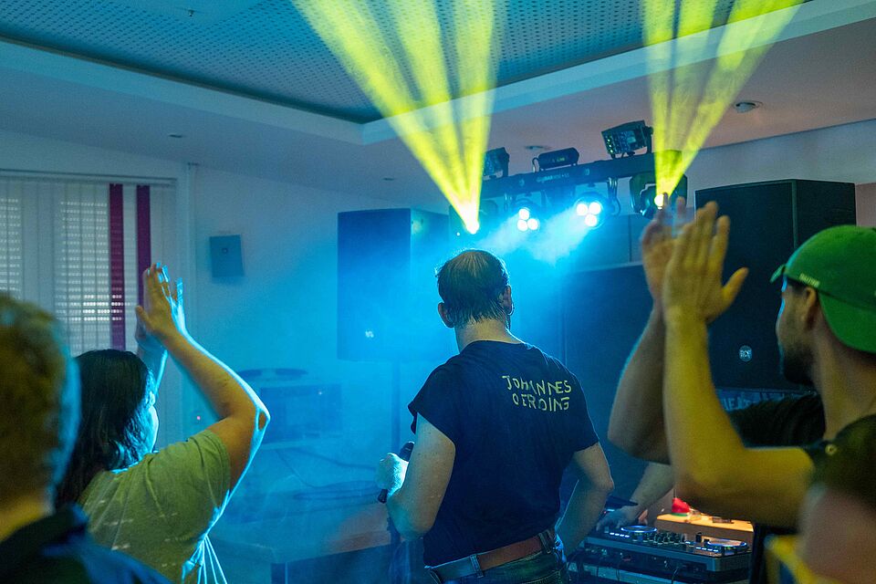 Das Bild zeigt eine Diskoveranstaltung mit bunten Lichteffekten, bei der ein DJ Musik auflegt und Feiernde tanzen.