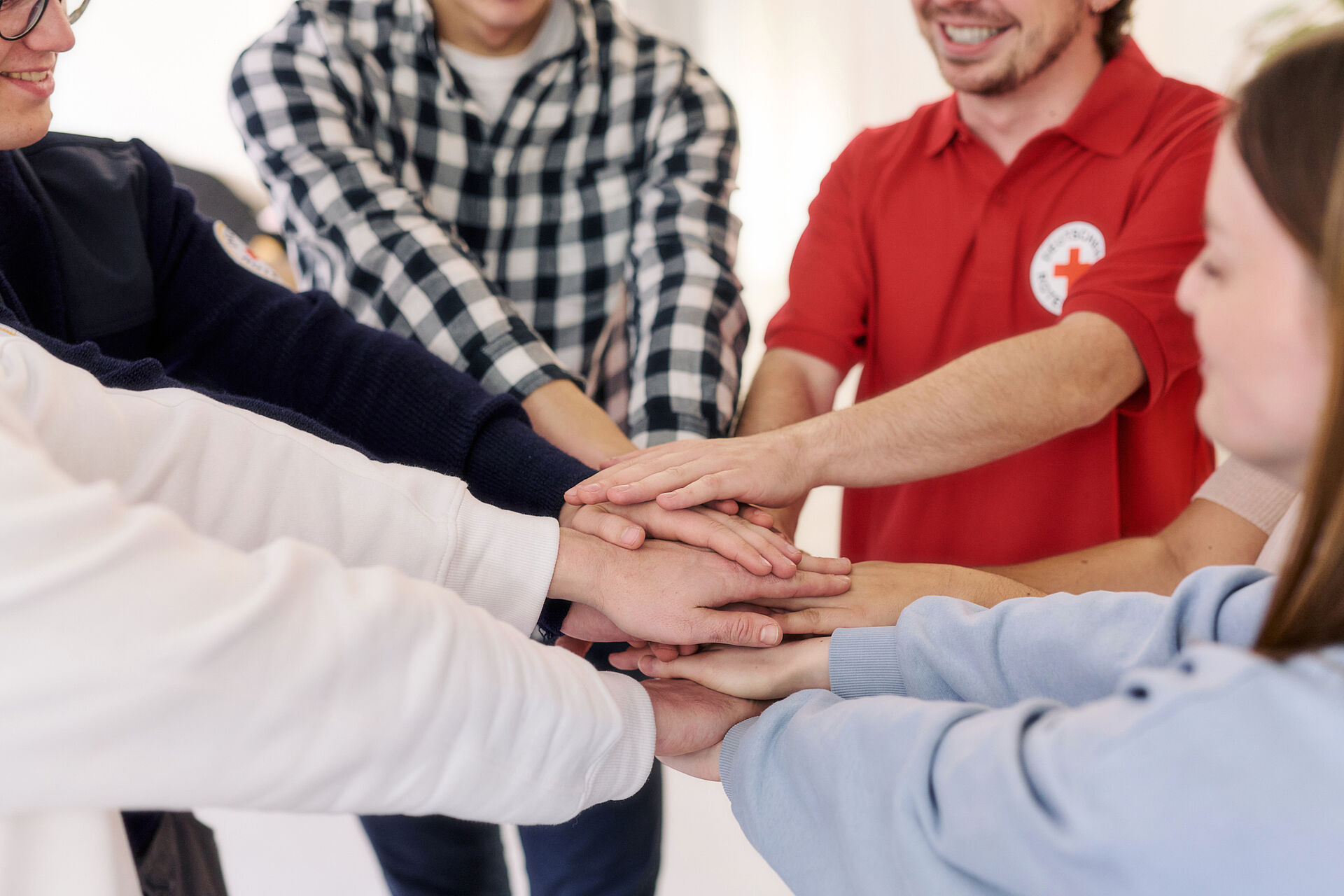 Eine Gruppe von Menschen, darunter ein Mann in einem roten DRK-Poloshirt, legt ihre Hände übereinander als Zeichen des Zusammenhalts.
