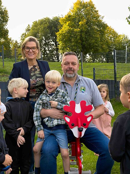 Homepage Waldwichtel Erwachsener sitzt mit einem Jungen auf einem roten Reh-Federwild-Wipptier im Grünen, dahinter steht eine Frau, weitere Kinder umgeben sie.