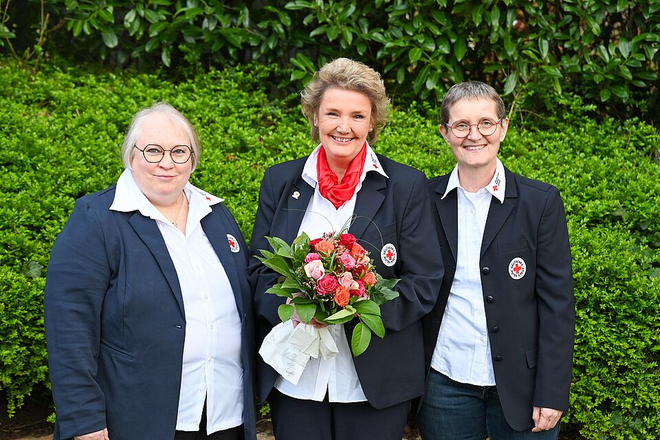 Drei Personen in Uniformen des Deutschen Roten Kreuzes stehen lächelnd vor einem grünen Buschhintergrund. Die mittlere Person hält einen Blumenstrauß mit roten und rosafarbenen Rosen. Alle tragen weiße Hemden mit dem DRK-Emblem und dunkle Jacken.