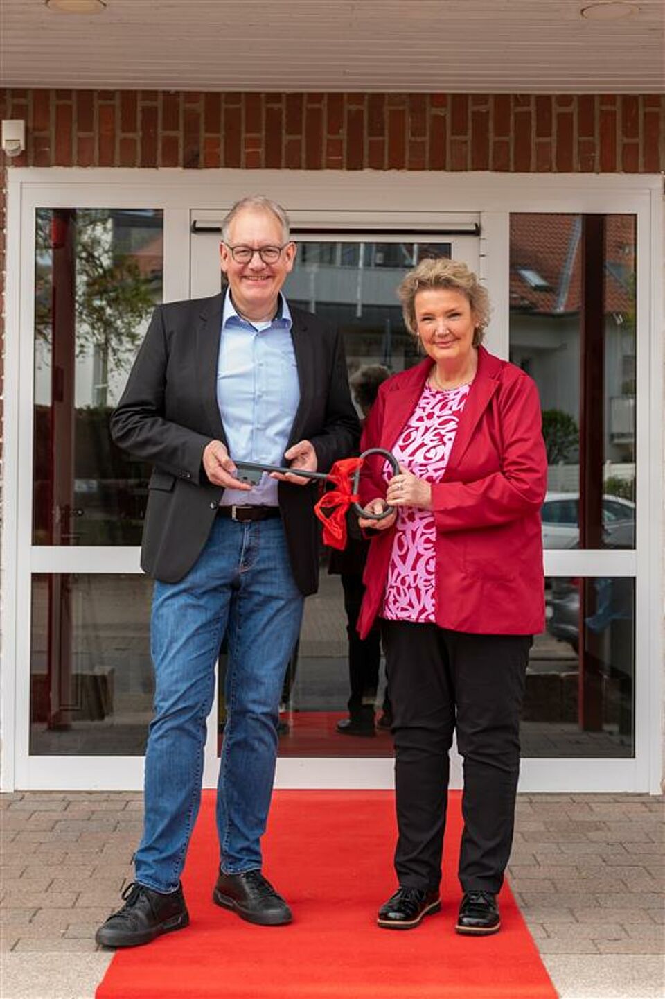 Zwei Personen stehen lächelnd auf einem roten Teppich vor einem Gebäude mit Glas- und Metalltüren. Die Person links trägt ein hellblaues Hemd und eine schwarze Jacke, die Person rechts ein rotes Jackett über einem weißen Oberteil. Beide halten gemeinsam eine rote Schleife und ein Glasobjekt als Symbol für eine Eröffnung oder Einweihung.