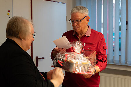 Das Foto zeigt eine freundliche, persönliche Übergabesituation in einem Innenraum (vermutlich Büro oder Gemeinschaftsraum). Ein älterer Mann mit grauen Haaren und Brille trägt ein rotes Oberteil und hält einen liebevoll verpackten Geschenkkorb in den Händen. Der Geschenkkorb ist in transparenter Folie eingeschlagen und mit roten Schleifen dekoriert. Zusätzlich liest der Mann eine Karte, die offenbar zum Geschenk gehört.  Ihm gegenüber steht eine ältere Frau mit Brille und kurzen grauen Haaren, die dunkel gekleidet ist. Sie lächelt und scheint das Geschenk gerade zu überreichen oder zu kommentieren. Die Körpersprache beider Personen wirkt wertschätzend und vertraut.