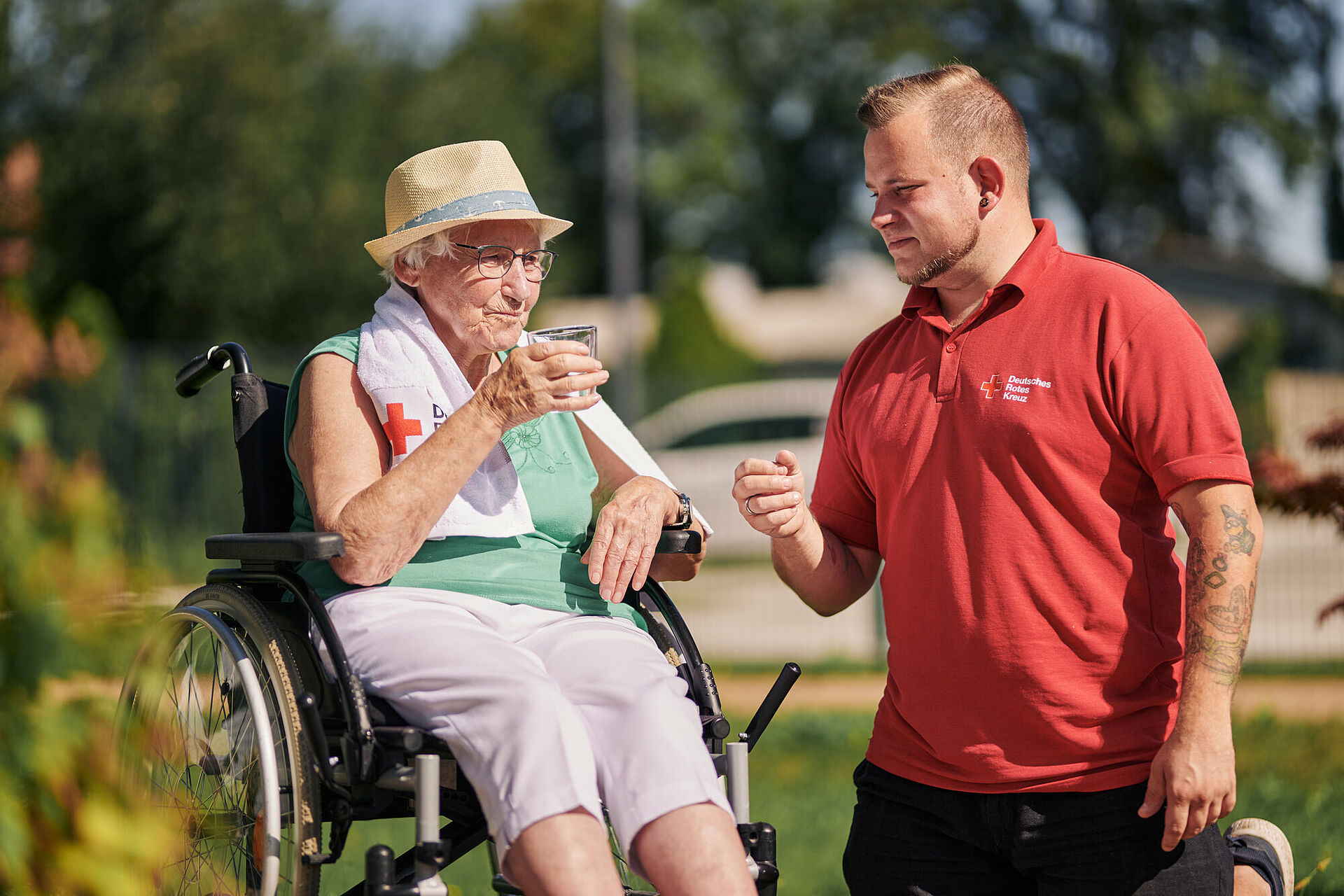 Ein Mann im roten DRK-Poloshirt kniet neben einer älteren Frau im Rollstuhl, die einen Hut trägt und aus einem Glas trinkt.