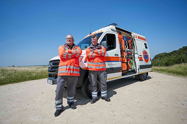 Zwei DRK-Einsatzkräfte in orangefarbenen Jacken stehen vor einem Einsatzwagen auf einem Feldweg.