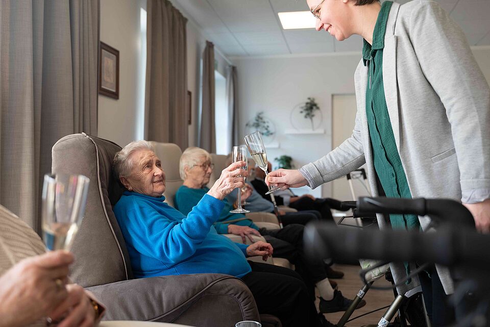 Mitarbeiterin stößt mit einer Seniorin in der DRK-Tagespflege mit einem Glas Sekt an, während weitere Gäste entspannt im Hintergrund sitzen.