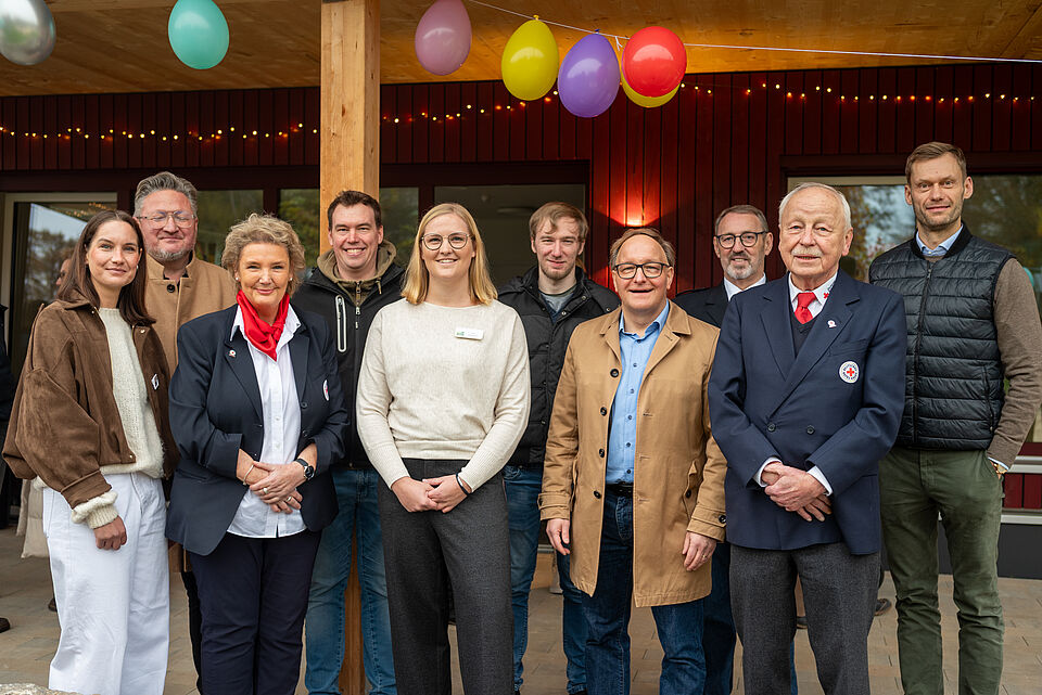 Eine Gruppe von zehn Personen steht lächelnd vor einem Holzgebäude mit bunter Luftballon-Dekoration. Es handelt sich um eine gemischte Gruppe aus Frauen und Männern unterschiedlichen Alters, die offenbar an einer feierlichen Veranstaltung oder Einweihung teilnehmen. Im Vordergrund stehen zwei Personen in dunkelblauer Kleidung mit roten Halstüchern, die an das Deutsche Rote Kreuz erinnern. Die übrigen tragen überwiegend helle oder braune Jacken und Pullover. Im Hintergrund sind rote Holzverkleidung, Lichterketten und Fenster zu sehen. Die Stimmung wirkt freundlich und festlich.